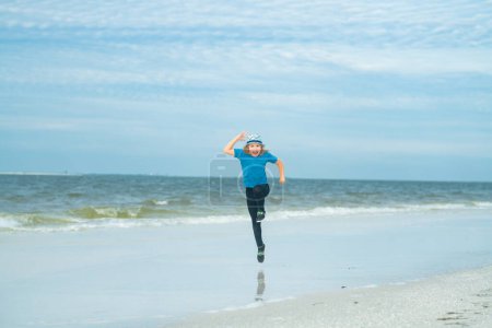 Foto de Lindo niño feliz corriendo a lo largo de la costa del mar al aire libre en la playa de verano. Un niño pequeño corre en la playa de verano. Un niño excitado corre contra el cielo azul y el mar. Concepto vacaciones de verano. Los niños juegan en el mar - Imagen libre de derechos