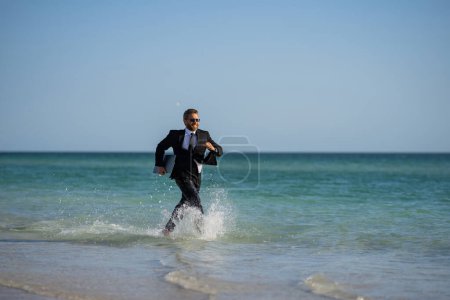 Téléchargez les photos : Homme d'affaires excité en combinaison humide courir en mer. Homme d'affaires drôle, concept d'affaires comique fou. Travail en ligne à distance. Des affaires d'été folles. Style de vie d'affaires amusant. Homme d'affaires freelance drôle - en image libre de droit