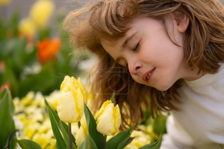 Foto de Lindo niño rubio bonito con los ojos cerrados oliendo flor de tulipán en el parque de primavera. Lindo niño pequeño sostiene tulipanes flores naturaleza. Retrato de niño en jardín de primavera con tulipanes - Imagen libre de derechos
