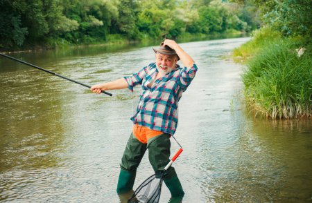 Foto de Emocionado pescador senior hombre con caña de pescar, carrete giratorio en el río. Viejo captura de peces, tirando de la caña mientras pesca en el lago - Imagen libre de derechos