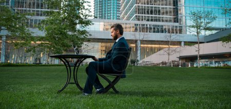 Téléchargez les photos : Homme d'affaires sur grand champ d'herbe près de l'immeuble de bureaux moderne. Homme d'affaires avec ordinateur portable dans le parc de mégapolice - en image libre de droit
