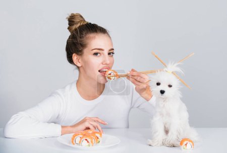 Foto de Young woman hold in hand makizushi sushi roll, traditional japanese food isolated background studio portrait. Woman and puppy dog with chopsticks eating sushi - Imagen libre de derechos
