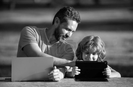 Téléchargez les photos : Père heureux utilisant ordinateur portable se détendre avec le fils écolier tenant ordinateur portable s'amuser ensemble, souriant papa et petit garçon enfant profiter week-end avec des gadgets à l'extérieur sur la nature. L'apprentissage en ligne - en image libre de droit