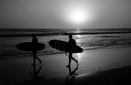 Téléchargez les photos : Silhouette de surfeurs portant leur planche de surf sur la plage du coucher du soleil - en image libre de droit