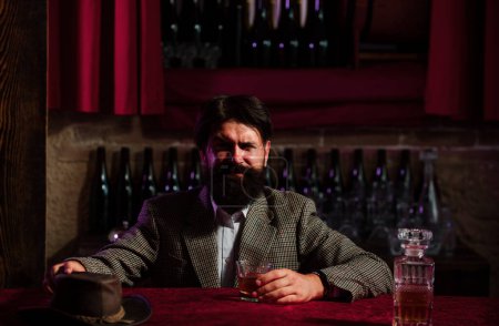 Téléchargez les photos : L'homme à la barbe tient du brandy en verre, boit du cognac dans un pub. Le mec hipster a le goût de boire. Siroter du whisky. Vie nocturne de luxe, riche homme barbu boisson chère - en image libre de droit