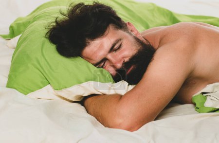 Foto de Foto de hombre guapo durmiendo y sosteniendo almohada verde suave. Hombre barbudo durmiendo en la cama en el dormitorio - Imagen libre de derechos
