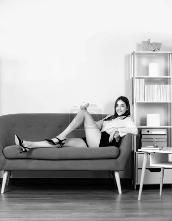 Secretaria seductora con piernas sexy sentarse en el sofá en la oficina. Mujer sexy de negocios bebiendo café. Chica sensual empleado