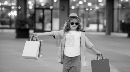 Foto de Lindo niño en ropa de moda de verano va de compras. Niño feliz con paquetes de compras en las manos. Shopper niño con bolsa de compras al aire libre. Lindo niño caminando en la calle llevando bolsas de compras - Imagen libre de derechos