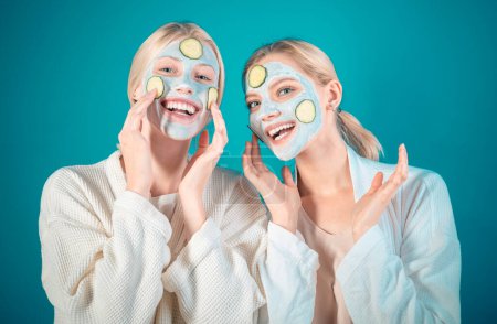Young happy sisters do facial mask with slices of cucumber at blue background. Skin care and treatment, spa, natural beauty and cosmetology concept