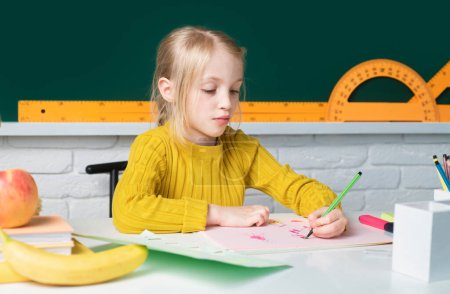 Photo for Portrait of cute little schoolgirl writing in book with classmates in background at classroom. Girl studying in school. Education, learning and children - Royalty Free Image