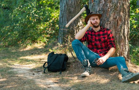 Téléchargez les photos : Ouvrier bûcheron assis dans la forêt. Bûcheron dans les bois avec une hache. Bûcheron avec hache à la main. Journalisation. Homme barbu avec concept de hache. Bûcheron tenant la hache - en image libre de droit