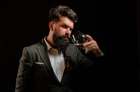Téléchargez les photos : Concept d'alcool. Élégant homme barbu élégant Barman tient verre de cognac. Homme ou homme d'affaires boit du cognac sur fond noir - en image libre de droit