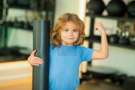 Téléchargez les photos : Enfant sportif montrant biceps tenant tapis de yoga dans la salle de gym. Yoga concept d'enfant. Jeune enfant fort et sportif montrant des biceps. Concept de sport d'entraînement - en image libre de droit