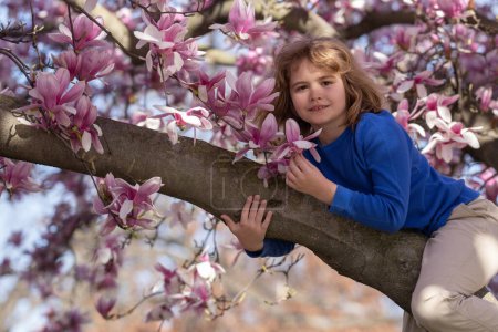 Froher Frühling. Kind klettert Magnolienbaum. Nahaufnahme Porträt eines lächelnden Kindergesichts in der Nähe blühender Frühlingsblumen. Kind zwischen Zweigen von Frühlingsbaum in Blüten