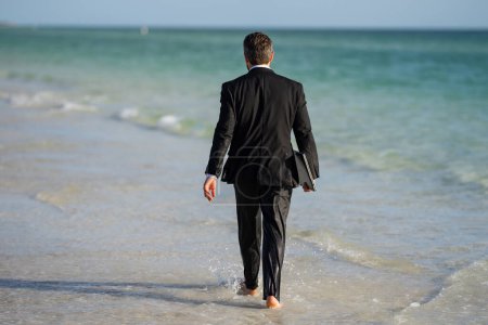 Téléchargez les photos : Homme d'affaires touriste en costume décontracté pieds nus marchant sur la plage tropicale avec ordinateur portable. Beau homme d'affaires en vacances d'été. Vue arrière du dos homme d'affaires en costume dans l'eau de mer à la plage - en image libre de droit