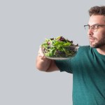Man hold fresh tomato and vegetable salad isolated on gray background, studio portrait. The vegetable greens. Healthy food. Portrait of handsome man hold vegetable near his face