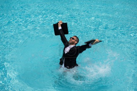 Téléchargez les photos : Homme d'affaires prospère en costume dans l'eau de piscine. Vacances d'été et concept de voyage. Homme d'affaires drôle en costume nager dans la piscine et tenant l'ordinateur portable. Humour, drôle concept d'entreprise d'été - en image libre de droit