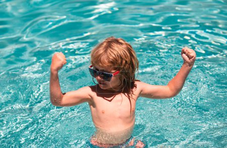 Téléchargez les photos : Mignon enfant garçon montrer les muscles, nager dans la piscine, fond d'eau d'été avec espace de copie. Visage drôle d'enfants - en image libre de droit