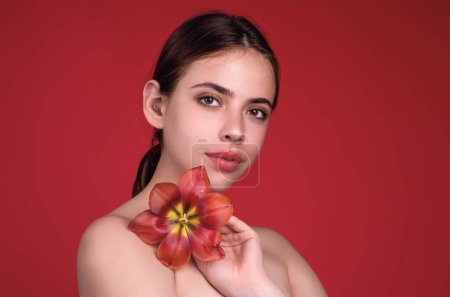 Foto de Ternura y belleza. Chica de belleza con tulipán cerca del hombro desnudo. Hermosa mujer sensual mantenga tulipanes, retrato de estudio sobre fondo rojo - Imagen libre de derechos