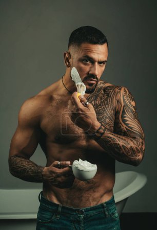 Téléchargez les photos : Bearded handsome tattooed man apply a shaving foam at his face. Bath procedures and grooming. Brutal muscular man going to shave his beard. Man style and fashion concept - en image libre de droit