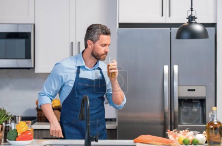 Foto de Happy man in cook apron cooking with bottle olive oil in kitchen. Portrait of middle aged man in chef apron in kitchen. Millennial man preparing food with olive oil - Imagen libre de derechos