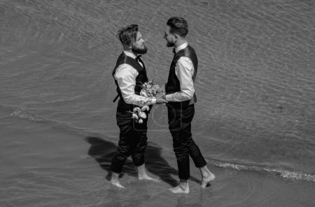 Photo for Gay grooms walking together on sea beach during Wedding day. Romantic men in sea water. Happy gay couple on wedding. Romantic and sensual gay couple. Gay grooms together on Wedding day - Royalty Free Image