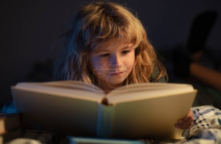 Foto de Retrato de primer plano del niño lindo leyendo un libro acostado en la cama. Niño leyendo un libro en la noche a la hora de dormir - Imagen libre de derechos