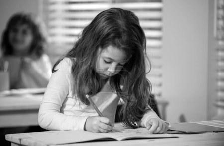 Téléchargez les photos : Écolière concentrée assise au bureau et écrivant dans un cahier d'exercices avec un camarade assis derrière - en image libre de droit