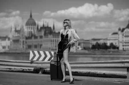 Foto de Turista sexy mujer en Budapest. Mujer turista en vestido de moda con maleta caminando por la calle. Chica estudiante viajero retrato al aire libre. Turista mujer teniendo viaje de vacaciones - Imagen libre de derechos