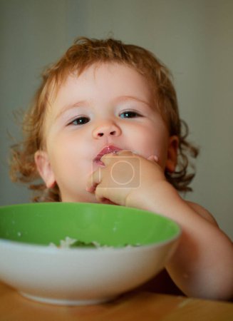 Téléchargez les photos : Bébé drôle mangeant lui-même avec une cuillère sur la cuisine. Enfant drôle visage gros plan - en image libre de droit