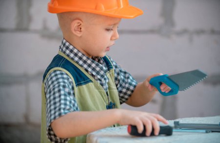 Téléchargez les photos : Garçon d'âge préscolaire essayant de voir, enfant sciant sur un chantier près du mur de la maison. Enfant blond avec casque utilise scie - en image libre de droit