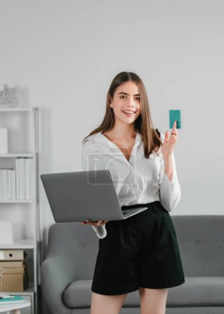 Photo for Young attractive secretary woman in a busy modern workplace in office. Pretty accountant girl with laptop and credit card at desk in office interior - Royalty Free Image