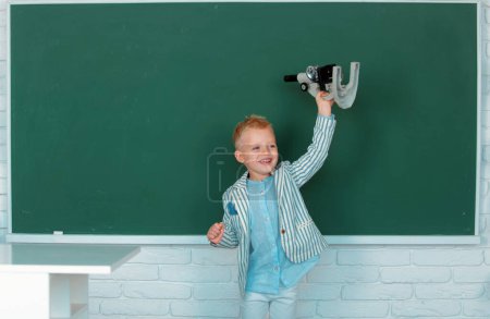 Photo for Child at school. Kid is learning in class on background of blackboard. Early education. Little child with microscope - Royalty Free Image
