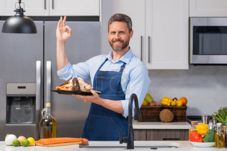 Photo for Happy man in cook apron cooking seafood in kitchen. Portrait of middle aged man in chef apron cooking seafood in kitchen. Millennial man preparing seafood - Royalty Free Image