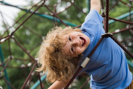 Téléchargez les photos : L'aire de jeux extérieure pour les enfants dans le parc d'été. Le gamin joue sur le terrain de jeu sous l'arbre. Portrait de jeune blonde excitée faisant de l'escalade avec de la verdure en arrière-plan - en image libre de droit