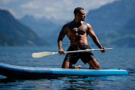 Sexy man paddling on paddleboard. Healthy summer lifestyle. Summer sport. SUP surfing tour in adventure summer vacation in Alps lake in Swiss. Male fit muscular model swimming with paddle board