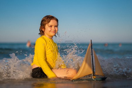 Photo for Little kid boy enjoying summer vacation by the sea. A cute little boy playing with a toy ship on the beach on a warm sunny summer day. Summer Holidays at sea. Kid playing with toy sailing boat - Royalty Free Image
