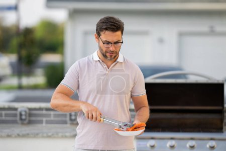 Photo for Barbecue master. Middle aged hispanic man in apron for barbecue. Roasting and grilling. Man hold cooking utensils barbecue. Roasting meat outdoors. Barbecue and grill. Cooking meat in home yard - Royalty Free Image