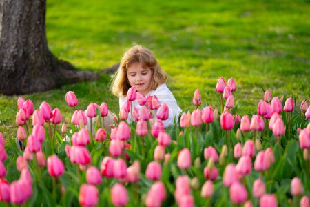 Foto de El niño sostiene la flor rosa del tulipán. Niño en el jardín de tulipanes. Niño jugar en el campo de tulipanes florecientes en el día de primavera. Cara de niños de primavera. Niño oliendo tulipán en el parque de primavera. Concepto primavera niños - Imagen libre de derechos