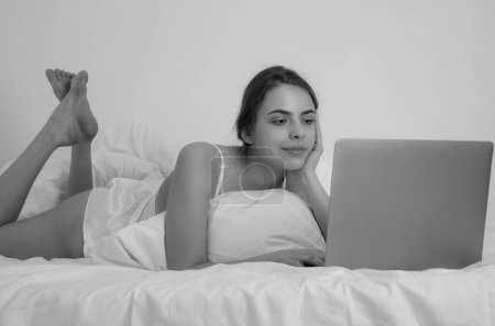 Téléchargez les photos : Woman watching laptop. Beautiful young woman using laptop on bed at home. Lazy freelancer girl with laptop sitting on bed. Girl work on laptop at morning resting in bed - en image libre de droit