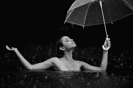 Téléchargez les photos : Femme avec parasol dans l'eau de mer du lac. Pluie estivale. Météo pluvieuse. La pluie s'en va. Pluie en été. Femme sexy sensuellement relaxant dans la piscine. Loisirs bien être et bien-être. Humide corps nu - en image libre de droit