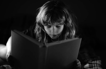 Téléchargez les photos : Enfant petit garçon lisant un livre dans le noir. Visage d'enfant avec veilleuse. Concept d'éducation et de lecture - en image libre de droit
