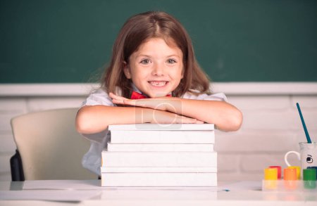 Téléchargez les photos : Fille souriante assise au bureau et tenant la main sur les livres dans la classe de l'école. Processus éducatif - en image libre de droit