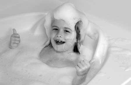 Foto de Cara de niños en espuma. Lavado de niños con burbujas en el baño. Lindo niño se baña, acostado en un baño blanco con espuma de jabón y champú - Imagen libre de derechos