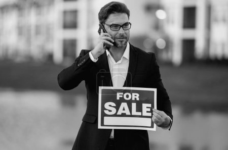 Photo for Real estate agent hold american house sale sign. Real estate sale or property investment concept. Buying new home. Handsome real estate agent in suit showing the house for sale - Royalty Free Image