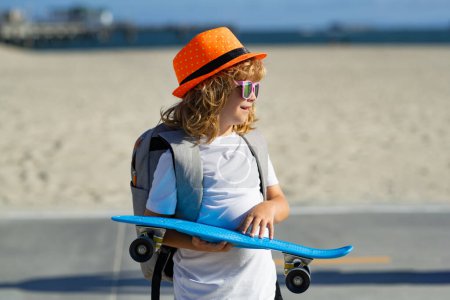 Téléchargez les photos : Portrait de mode en plein air de l'enfant élégant dans le chapeau de mode, lunettes de soleil de mode tiennent skateboard près de la rue. Style de vie des enfants urbains extérieurs - en image libre de droit