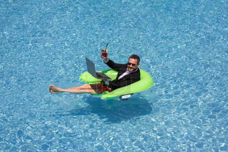 Téléchargez les photos : Vacances d'été et concept de voyage de liberté. Portrait d'un homme d'affaires hispanique sur la plage. Homme d'affaires en costume flottant avec ordinateur portable et cocktail dans la piscine. Homme d'affaires fou rêve vacances d'été - en image libre de droit