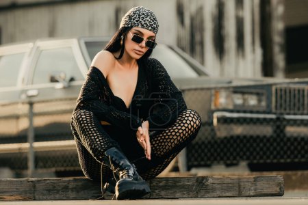 Photo for Pretty sexy fashion sensual woman posing on garage wall. Hipster young woman in hipster style outfit. Stylish fashionable girl in black clothes. Beautiful sexy woman sitting near retro car - Royalty Free Image
