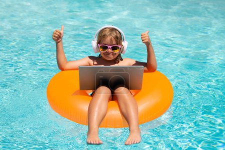 Téléchargez les photos : Un petit homme d'affaires qui travaille sur une plage tropicale. Enfant relaxant dans la piscine avec ordinateur portable. Enfant travaillant en ligne sur ordinateur portable, nageant dans une piscine d'eau turquoise ensoleillée - en image libre de droit
