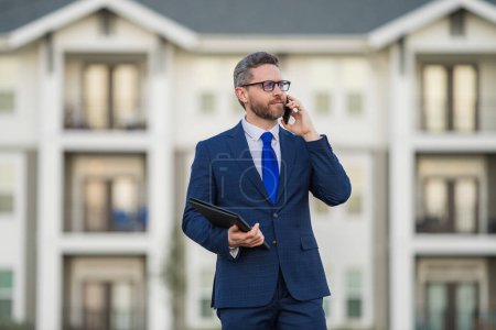 Photo for Real estate agent business man in suit hold holder clipboards and talking on phone. Businessman. Business Man broker or real estate agent. Business man in suit outdoor. Real estate business man - Royalty Free Image